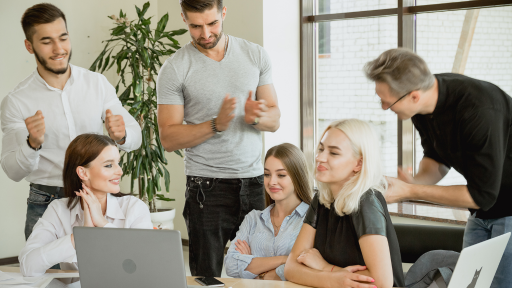 A Imagem 3 homens em pé, cantam parabéns a você para 3 mulheres sentadas diante de computador, o clima é descontraído. Image 3 men standing, singing happy birthday to 3 women sitting in front of a computer, the atmosphere is relaxed.