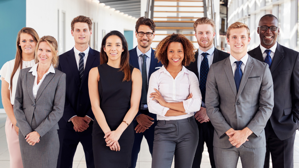 A Imagem mostra 9 pessoas de diversas etnias, sendo 5 homens vestindo terno e  4 mulheres, de diversas idades, estão 2 filas, ao fundo um ambiente coorporativo e uma escada para o andar superior.  The Image shows 9 people of different ethnicities, 5 men wearing suits and 4 women, of different ages, in 2 rows, in the background a corporate environment and a staircase to the upper floor.